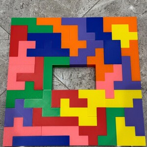 May include: A colourful wooden puzzle with a square frame and a hole in the centre. The puzzle pieces are arranged in a pattern of red, orange, yellow, green, blue, and pink.