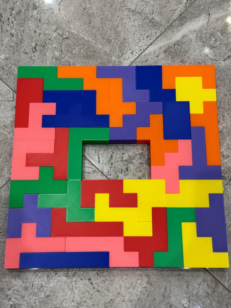 May include: A colourful wooden puzzle with a square frame and a hole in the centre. The puzzle pieces are arranged in a pattern of red, orange, yellow, green, blue, and pink.