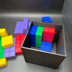 May include: A black square container holds a collection of colorful, geometric puzzle pieces. The pieces are various shapes and colors, including red, blue, green, yellow, and purple. Some pieces are inside the container, while others are scattered around it.