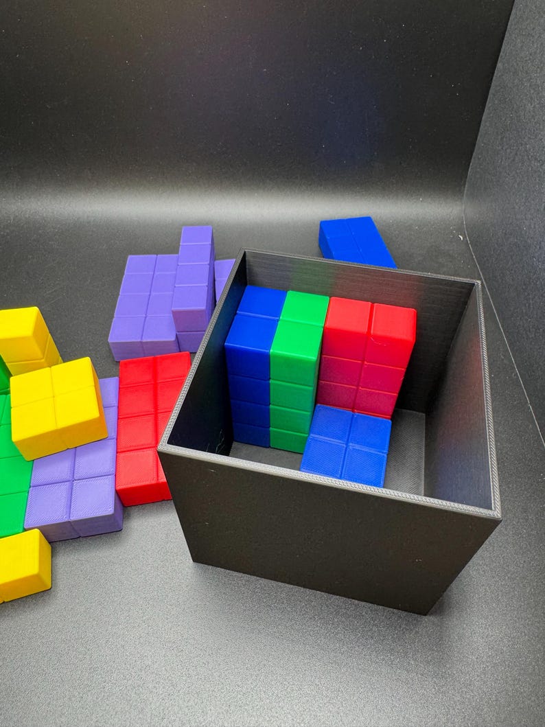 May include: A black square container holds a collection of colorful, geometric puzzle pieces. The pieces are various shapes and colors, including red, blue, green, yellow, and purple. Some pieces are inside the container, while others are scattered around it.