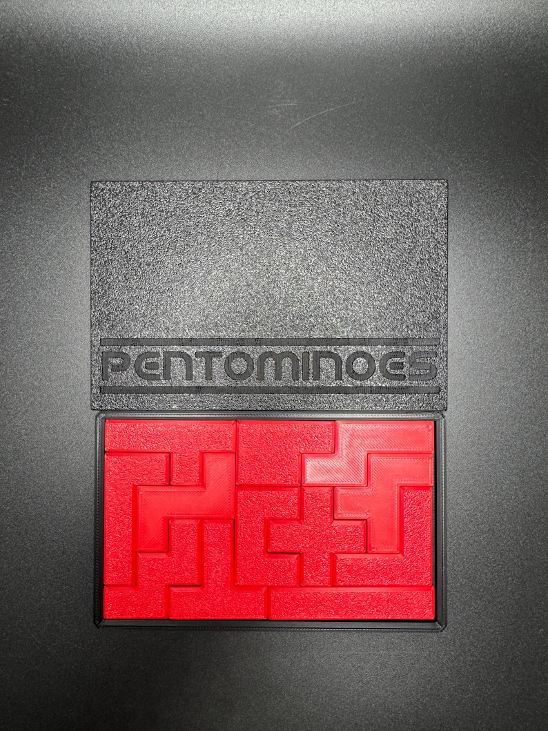 May include: A red plastic pentominoes puzzle set in a black plastic case with the word "PENTOMINOES" printed on the case.