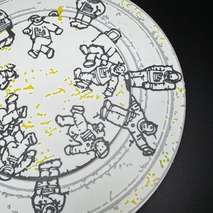 Puede incluir: Plato de cer&aacute;mica blanca con un borde gris y un dise&ntilde;o de astronautas en contornos negros. El plato tiene motas amarillas y un fondo negro. Los astronautas est&aacute;n en varias poses, dando al plato una est&eacute;tica l&uacute;dica con tem&aacute;tica espacial.