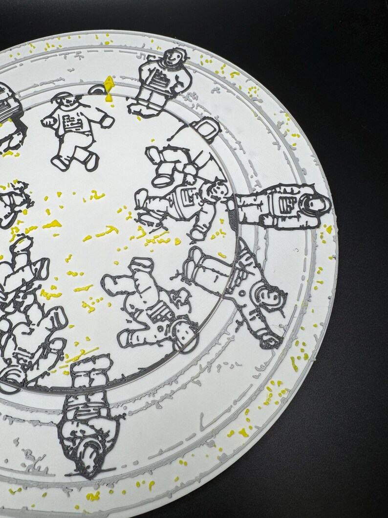Puede incluir: Plato de cer&aacute;mica blanca con un borde gris y un dise&ntilde;o de astronautas en contornos negros. El plato tiene motas amarillas y un fondo negro. Los astronautas est&aacute;n en varias poses, dando al plato una est&eacute;tica l&uacute;dica con tem&aacute;tica espacial.