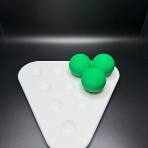 May include: A white triangular tray with seven circular indentations, three green balls are sitting on top of the tray.