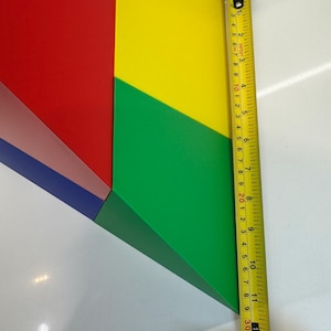 May include: A colorful geometric shape made of four pieces of cardboard. The pieces are red, yellow, green, and blue. The shape is measured with a tape measure, which shows a measurement of 11 inches.