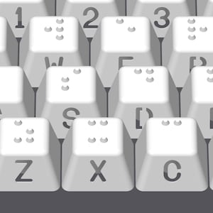 May include: A white keyboard with braille markings on the keys. The keys are labeled with letters and numbers. The keyboard is on a gray surface.