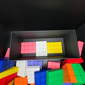 May include: A collection of colorful plastic building blocks in various colors, including red, yellow, blue, green, white, and pink. The blocks are arranged in a box and on a black surface.