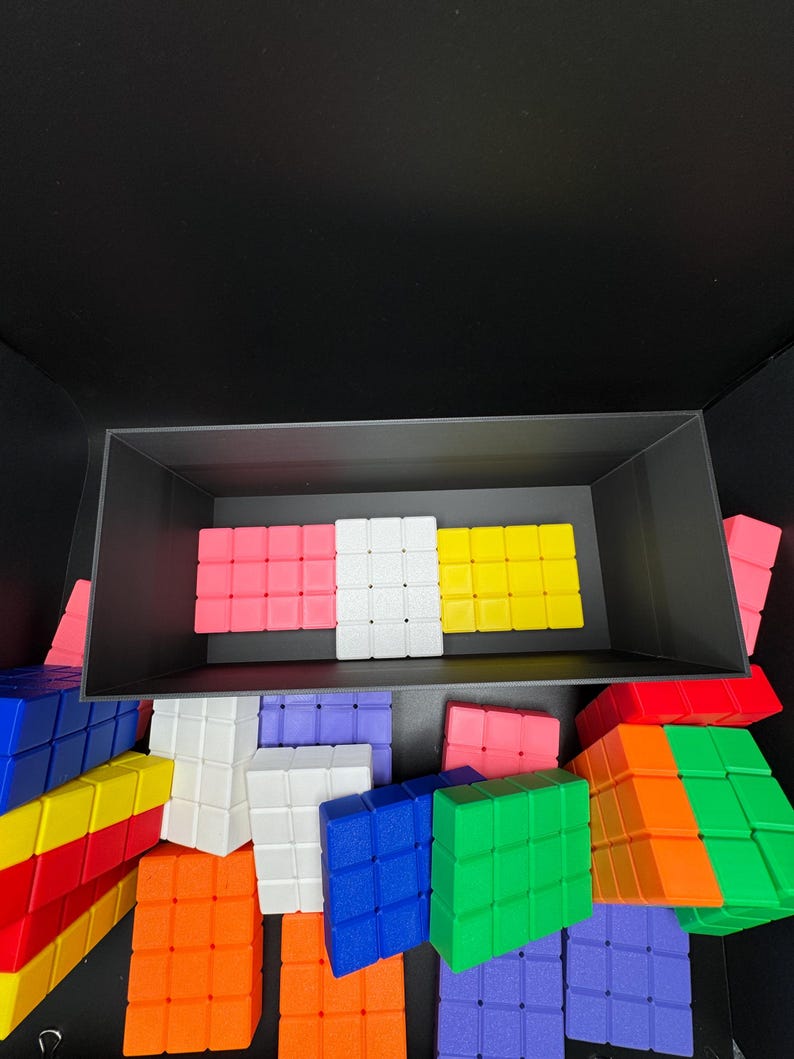 May include: A collection of colorful plastic building blocks in various colors, including red, yellow, blue, green, white, and pink. The blocks are arranged in a box and on a black surface.