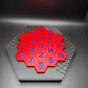Puede incluir: Un rompecabezas hexagonal rojo con n&uacute;meros azules del 1 al 19 en cada pieza. El rompecabezas est&aacute; colocado sobre una base hexagonal negra con un patr&oacute;n de panal.