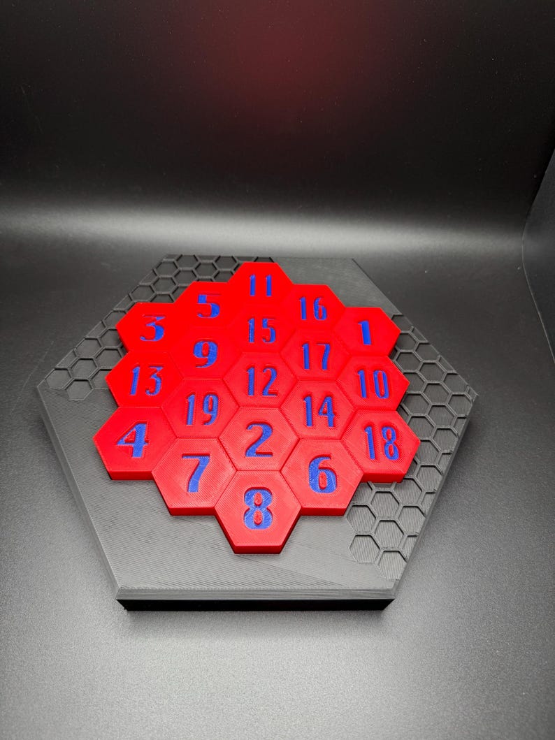 Puede incluir: Un rompecabezas hexagonal rojo con n&uacute;meros azules del 1 al 19 en cada pieza. El rompecabezas est&aacute; colocado sobre una base hexagonal negra con un patr&oacute;n de panal.