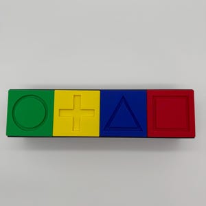 May include: A black rectangular block with four colored squares. The squares are green, yellow, blue, and red. Each square has a different shape embossed on it: a circle, a plus sign, a triangle, and a square.