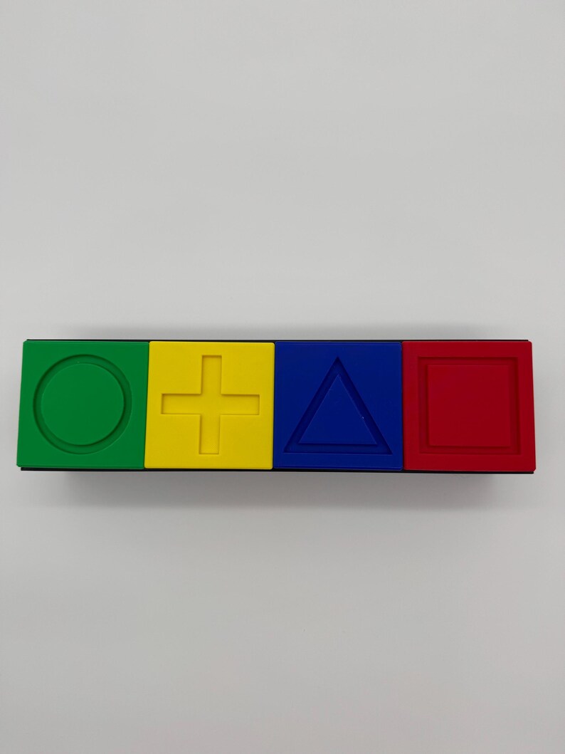 May include: A black rectangular block with four colored squares. The squares are green, yellow, blue, and red. Each square has a different shape embossed on it: a circle, a plus sign, a triangle, and a square.