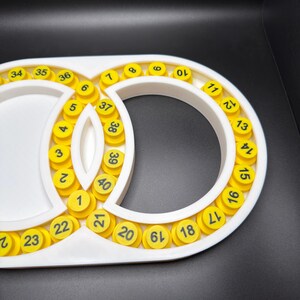 May include: A white plastic frame with a figure-eight shape containing forty circular yellow inserts. Each yellow insert is printed with a black number ranging from 1 to 40, arranged in a sequential pattern along the inner and outer edges.