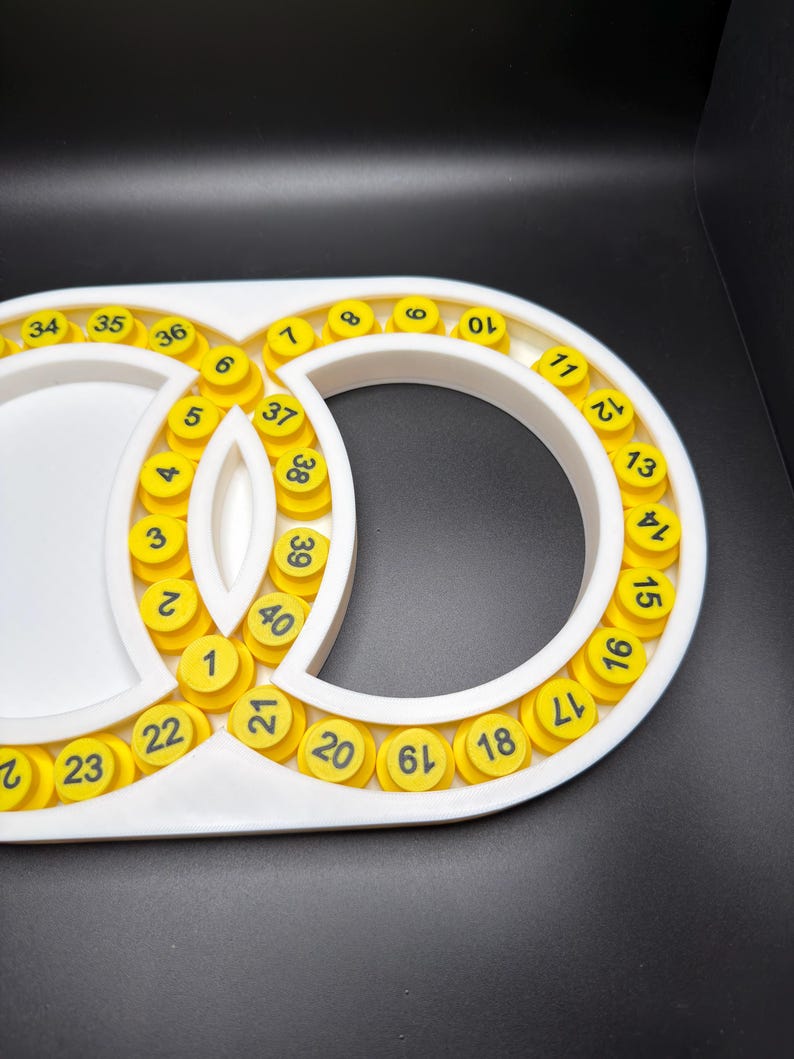 May include: A white plastic frame with a figure-eight shape containing forty circular yellow inserts. Each yellow insert is printed with a black number ranging from 1 to 40, arranged in a sequential pattern along the inner and outer edges.