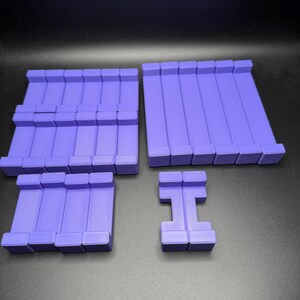 May include: A set of four purple plastic building blocks. The blocks are rectangular and have a smooth surface. They are arranged in a pattern on a gray surface.