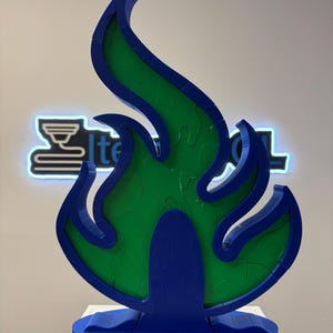 May include: A blue and green flame-shaped sculpture. The sculpture has a blue base and frame, with a green interior. The flame design is a puzzle. The sculpture is on a white surface, with a neon sign in the background.