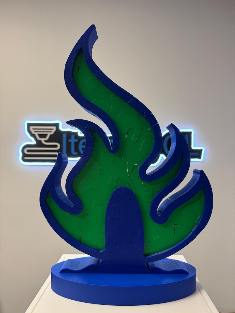 May include: A blue and green flame-shaped sculpture. The sculpture has a blue base and frame, with a green interior. The flame design is a puzzle. The sculpture is on a white surface, with a neon sign in the background.