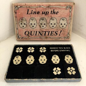 May include: A vintage game box with a pink lid and black interior. The lid features the text "Line up the QUINTIES!" with five baby faces. The box contains a game board with five baby faces and three flower tiles. The game board is black with white outlines. The game box is labeled "REMOVE THIS BLOCK BEFORE STARTING."