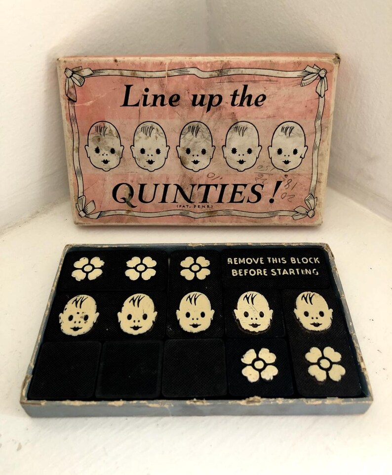 May include: A vintage game box with a pink lid and black interior. The lid features the text "Line up the QUINTIES!" with five baby faces. The box contains a game board with five baby faces and three flower tiles. The game board is black with white outlines. The game box is labeled "REMOVE THIS BLOCK BEFORE STARTING."