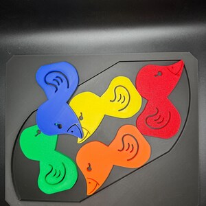 May include: A colorful puzzle featuring six bird-shaped pieces in blue, green, yellow, orange, and red. The pieces fit into a black, rectangular base with a curved outline. Each bird has a beak and an ear.