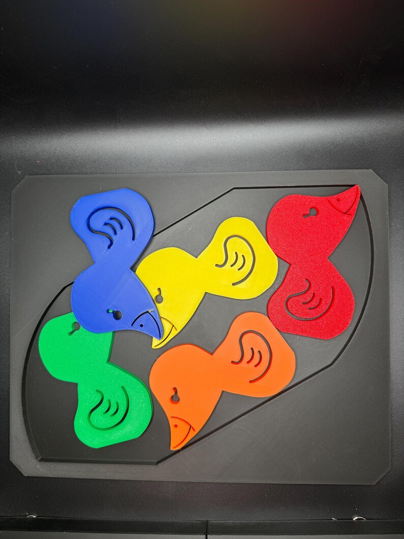 May include: A colorful puzzle featuring six bird-shaped pieces in blue, green, yellow, orange, and red. The pieces fit into a black, rectangular base with a curved outline. Each bird has a beak and an ear.