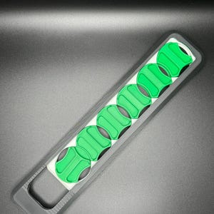 May include: A black and white plastic tool with a handle and eight green circular slots. The tool is designed to hold and organize small round objects.