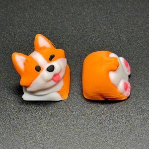 Op de afbeelding: Twee 3D-geprinte corgi-beeldjes. E&eacute;n is een vooraanzicht met oranje, witte en zwarte details, inclusief een roze tong. De andere toont de achterkant van de corgi, met roze voetzolen. De beeldjes staan op een grijze ondergrond.