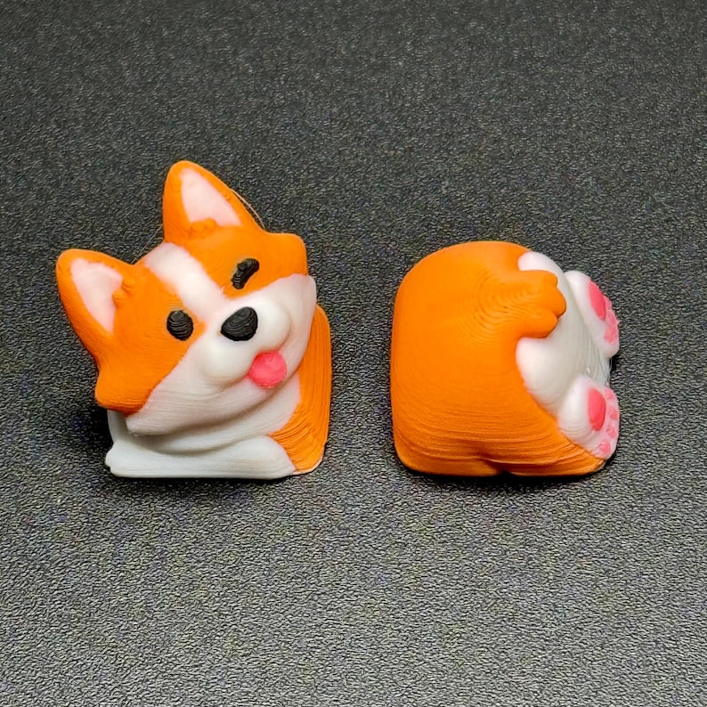 Op de afbeelding: Twee 3D-geprinte corgi-beeldjes. E&eacute;n is een vooraanzicht met oranje, witte en zwarte details, inclusief een roze tong. De andere toont de achterkant van de corgi, met roze voetzolen. De beeldjes staan op een grijze ondergrond.