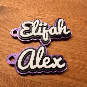 K&ouml;nnte beinhalten: Zwei violette, 3D-gedruckte Namensschilder mit wei&szlig;en Buchstaben. Das obere Schild sagt "Elijah" und das untere Schild sagt "Alex".