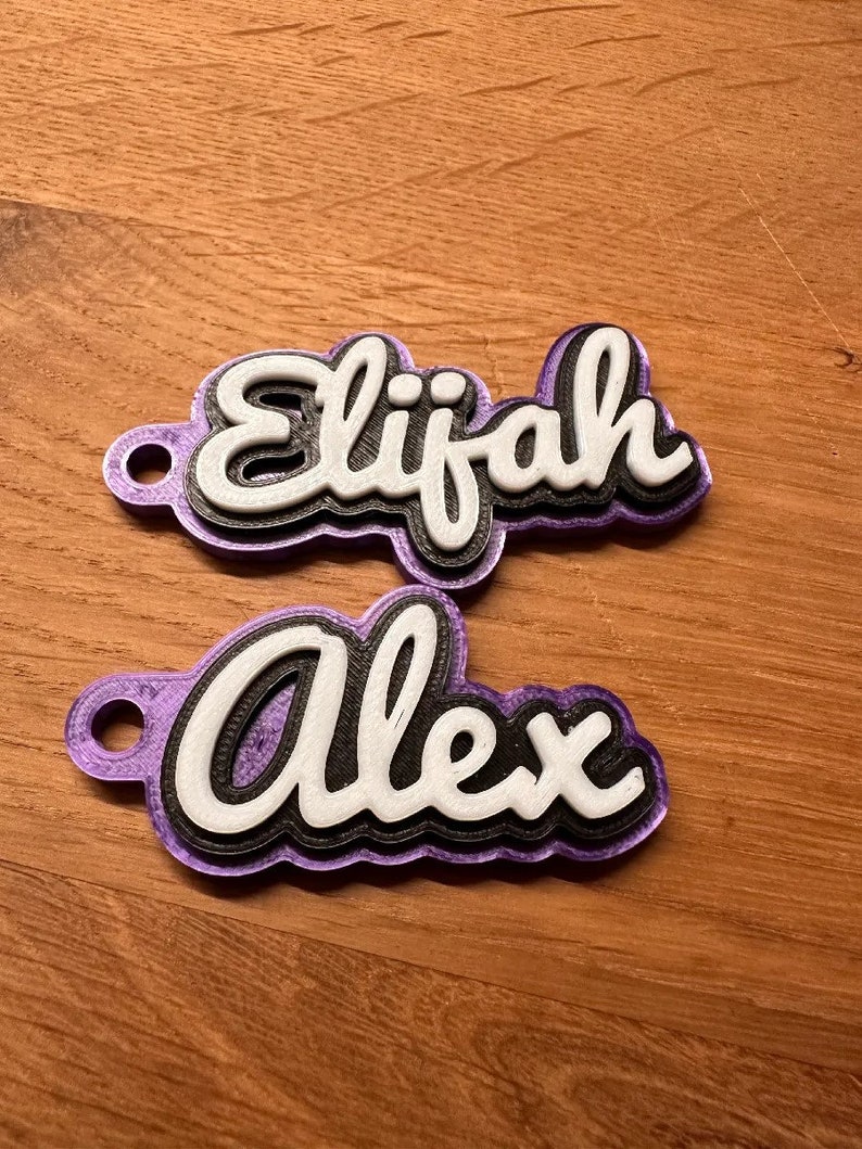 K&ouml;nnte beinhalten: Zwei violette, 3D-gedruckte Namensschilder mit wei&szlig;en Buchstaben. Das obere Schild sagt "Elijah" und das untere Schild sagt "Alex".