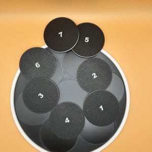 May include: A set of black circular coasters with white numbers 1 through 6 printed on them. The coasters are arranged on a larger, gray, circular base with a white border. The background is a solid orange color.