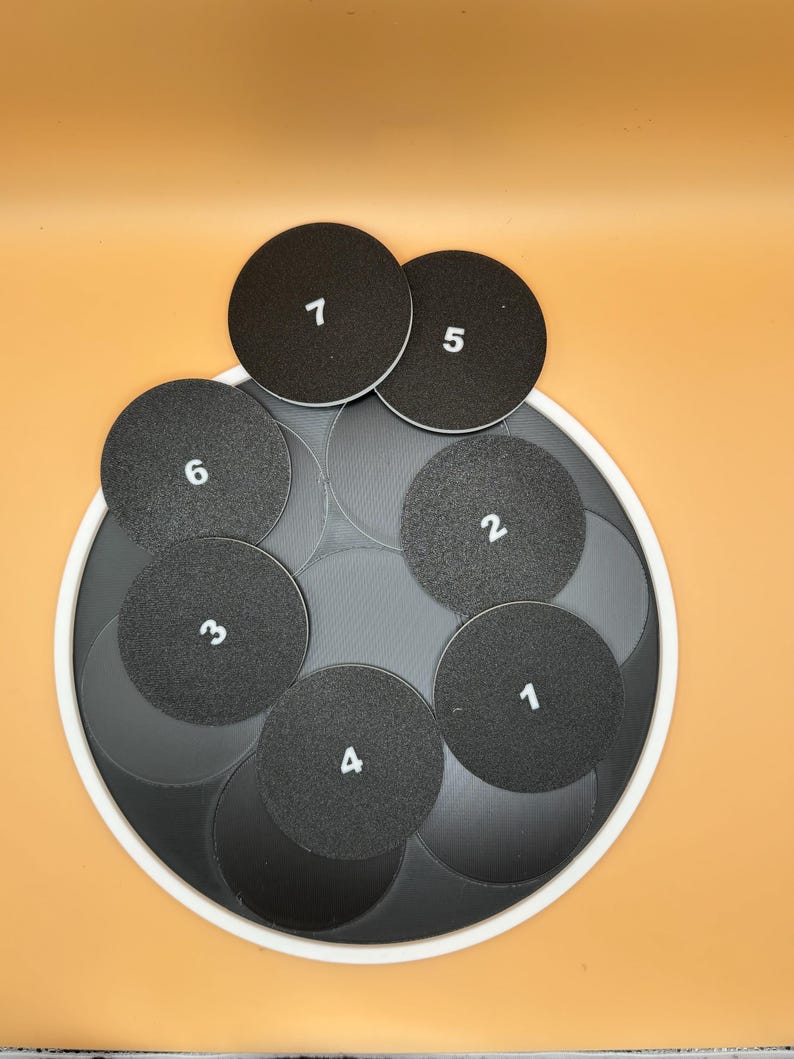 May include: A set of black circular coasters with white numbers 1 through 6 printed on them. The coasters are arranged on a larger, gray, circular base with a white border. The background is a solid orange color.