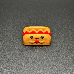May include: A small, 3D printed hot dog with a smiling face and a red heart in its hands. The hot dog is yellow with a red and yellow mustard topping.