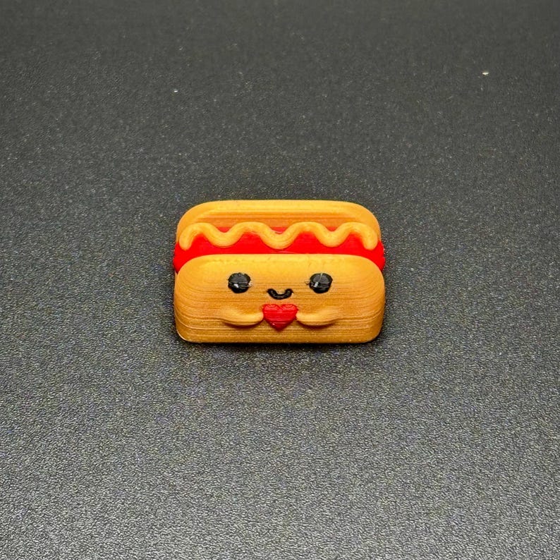 May include: A small, 3D printed hot dog with a smiling face and a red heart in its hands. The hot dog is yellow with a red and yellow mustard topping.