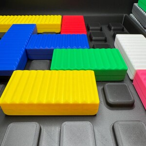 May include: A collection of colorful, rectangular, ridged blocks in yellow, blue, green, red, white, and pink. The blocks are arranged on a black surface with square indentations. The blocks appear to be part of a puzzle or building set.