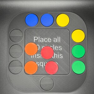 K&ouml;nnte beinhalten: Ein graues Puzzlespiel mit quadratischer Mitte und kreisf&ouml;rmigen Aussparungen. Es hat blaue, gelbe, orangefarbene, rote und gr&uuml;ne kreisf&ouml;rmige Teile. Der Text auf dem Brett lautet: "Alle Kreise in dieses Quadrat legen."