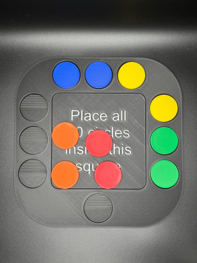 K&ouml;nnte beinhalten: Ein graues Puzzlespiel mit quadratischer Mitte und kreisf&ouml;rmigen Aussparungen. Es hat blaue, gelbe, orangefarbene, rote und gr&uuml;ne kreisf&ouml;rmige Teile. Der Text auf dem Brett lautet: "Alle Kreise in dieses Quadrat legen."