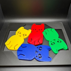 May include: A collection of five colorful cat-shaped cutouts in yellow, red, green, blue, and yellow. Each cat has a simple, stylized design with cut-out eyes and a curved tail. The cutouts are arranged on a dark gray surface.