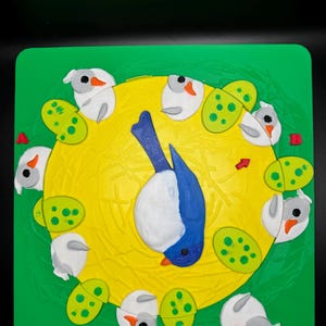 May include: A colorful, handcrafted puzzle featuring a blue and white bird in the center, surrounded by white birds and green, spotted shapes. The puzzle is set against a yellow circle and a green square background.