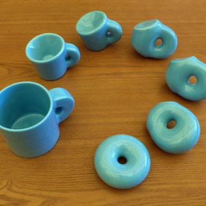 Op de afbeelding: Een verzameling lichtblauwe keramische mokken en donutvormige objecten op een houten oppervlak. De mokken hebben afgeronde handvatten. De donutvormige objecten hebben een gat in het midden. Alle objecten hebben dezelfde blauwtint.