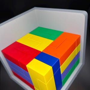 May include: A multi-colored 3D puzzle cube composed of interlocking plastic blocks in vibrant shades of red, orange, yellow, green, blue, and purple. The puzzle is arranged in a square formation and sits within a white, L-shaped corner display frame.