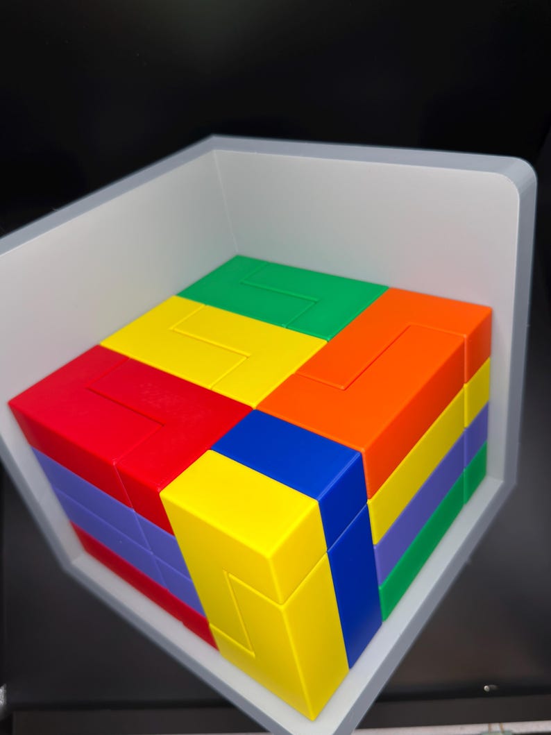 May include: A multi-colored 3D puzzle cube composed of interlocking plastic blocks in vibrant shades of red, orange, yellow, green, blue, and purple. The puzzle is arranged in a square formation and sits within a white, L-shaped corner display frame.