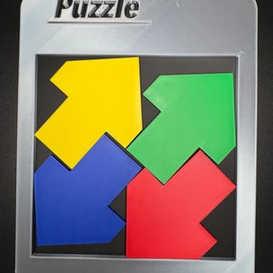 Pu&ograve; includere: Un puzzle colorato con quattro pezzi a forma di freccia gialli, verdi, blu e rossi. La parola "Puzzle" &egrave; in rilievo sulla cornice argentata. I pezzi si inseriscono in un quadrato nero, all'interno di una cornice rettangolare argentata.