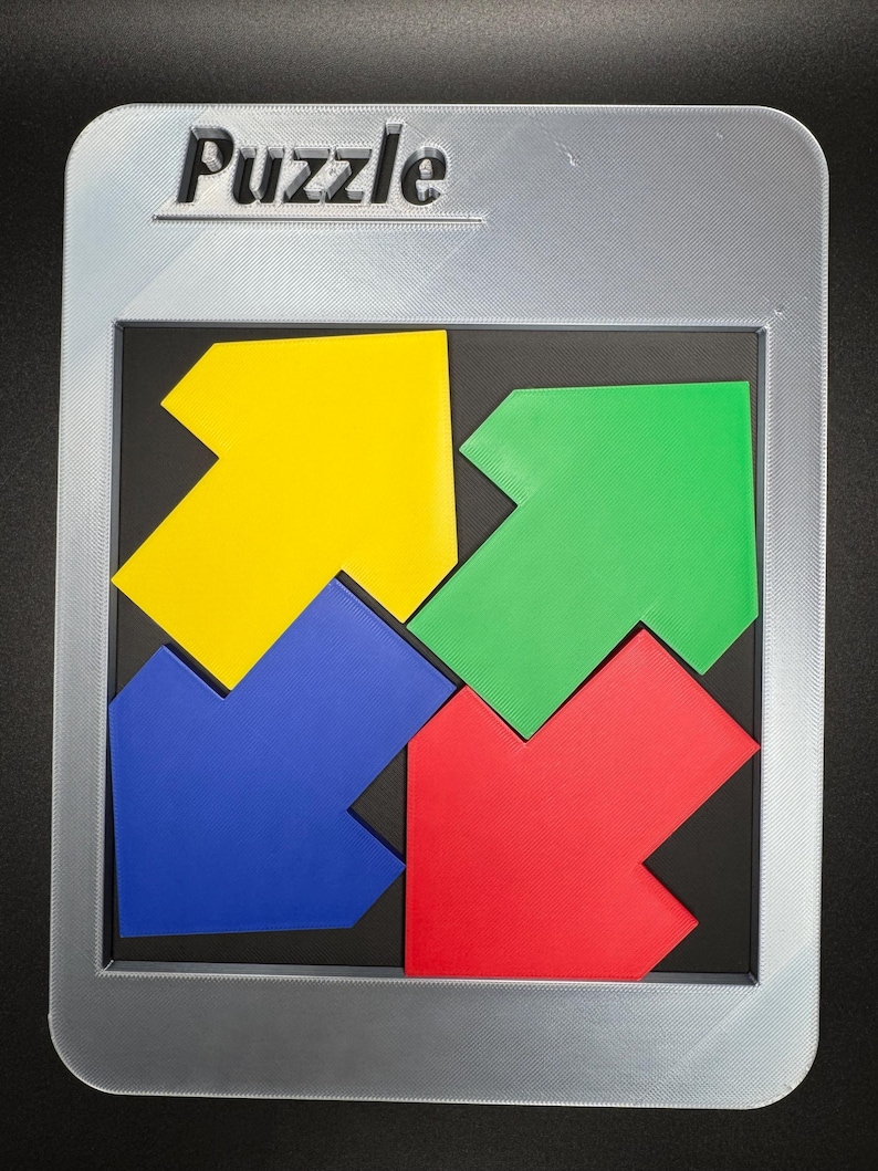 Pu&ograve; includere: Un puzzle colorato con quattro pezzi a forma di freccia gialli, verdi, blu e rossi. La parola "Puzzle" &egrave; in rilievo sulla cornice argentata. I pezzi si inseriscono in un quadrato nero, all'interno di una cornice rettangolare argentata.