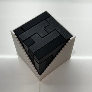 Puede incluir: Un rompecabezas 3D negro en forma de la letra 'H' est&aacute; ensamblado y colocado dentro de una base blanca, angulada y geom&eacute;trica.