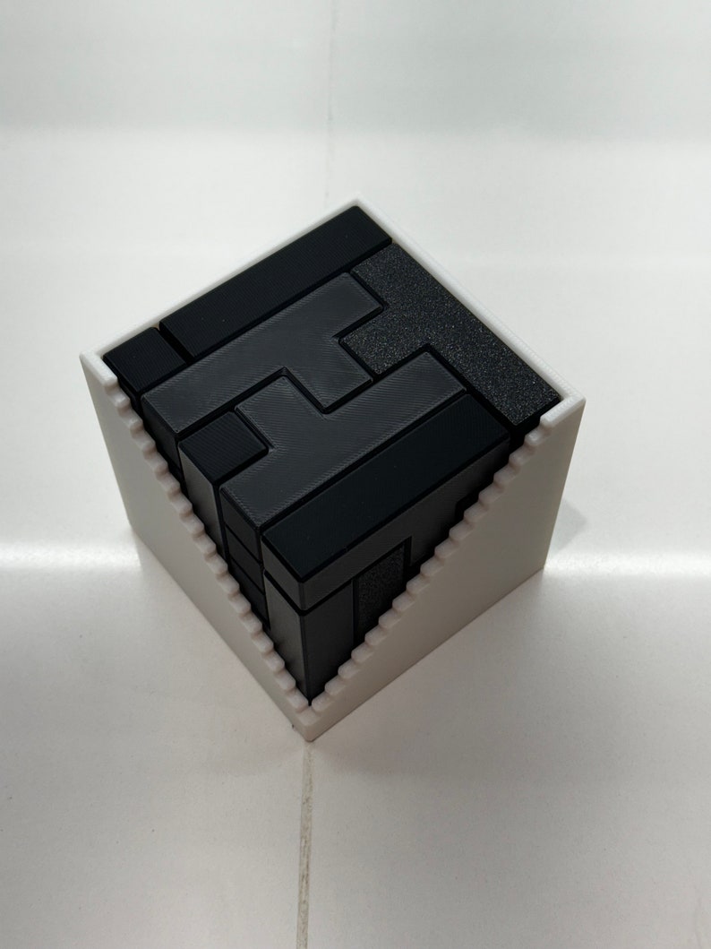 Puede incluir: Un rompecabezas 3D negro en forma de la letra 'H' est&aacute; ensamblado y colocado dentro de una base blanca, angulada y geom&eacute;trica.