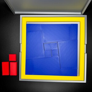 May include: A blue puzzle with a yellow frame, made up of smaller squares, is in a gray box with a yellow frame. Two red blocks are on the left side of the image.