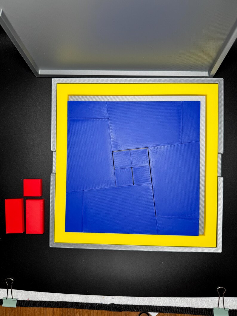 May include: A blue puzzle with a yellow frame, made up of smaller squares, is in a gray box with a yellow frame. Two red blocks are on the left side of the image.