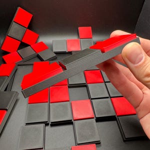 K&ouml;nnte beinhalten: Eine Sammlung roter und schwarzer geometrischer Puzzleteile. Die Teile sind auf einer dunklen Oberfl&auml;che angeordnet, einige werden in einer Hand gehalten. Die Puzzleteile sind quadratisch und rechteckig, mit einem kontrastierenden Farbschema.