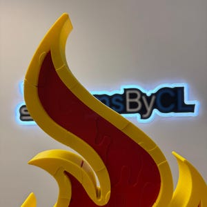May include: A close-up of a flame-shaped puzzle. The puzzle is primarily red with yellow edges. The puzzle pieces are interlocking. The background has the text "Signs By CL" in a neon blue glow.